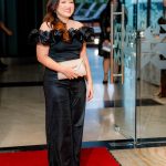Woman in a black off-shoulder outfit holds a clutch and stands on a red carpet indoors.