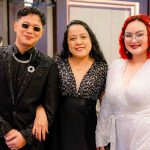 Three people dressed in formal attire pose together and smile at an indoor event.