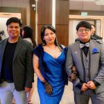 Three people dressed in formal attire pose together at an indoor event or party.