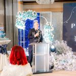 A man speaks at a podium during a formal indoor event with blue and white decorations.