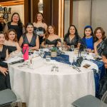 A group of women in formal attire seated around a round table at an indoor event.