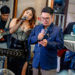 Man and woman singing with microphones at an event, guitarist playing in the background.