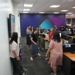 A group of people standing in an office, engaged in conversation and laughter near desks and computer monitors.
