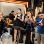 Four singers perform in formal attire alongside musicians at an indoor event.