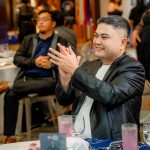 Man in a black jacket claps while seated at a formal event with drinks on the table.