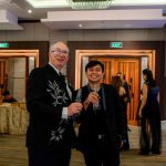 Two people in formal attire smile and hold drinks at an indoor event.