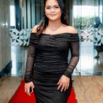 Woman in a black off-shoulder gown stands on a red carpet in a modern lobby.