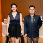 A woman in a black dress and a man in a suit stand indoors near a buffet table.