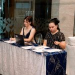 Two women sit at a registration table with papers, a laptop, and stacked plates nearby.