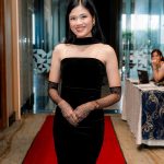 Woman in a black dress stands on a red carpet in a modern hallway, smiling at the camera.