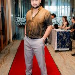 Man in gradient brown shirt and light pants stands on a red carpet in a hallway.