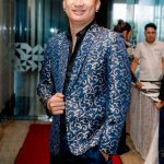 Man in a blue patterned blazer stands on a red carpet in a formal indoor setting.