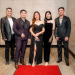 Five people in formal attire stand together on a red carpet in an indoor setting.