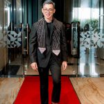 Man in a black suit with a patterned scarf stands on a red carpet in a modern hallway.