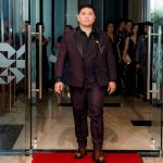 A man in a suit walks on a red carpet through glass doors at an indoor event.