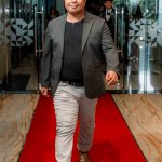 Man in a blazer and slacks walking confidently on a red carpet indoors.