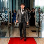 A man in a suit walks on a red carpet through glass doors at an indoor event.