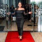 Woman in a black dress walking on a red carpet with glass doors and people in the background.
