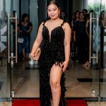 Woman in a black evening gown with a slit poses on a red carpet, holding a clutch.