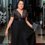 Woman in a black evening gown holding a clutch walks on a red carpet indoors.