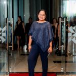 Person in a blue outfit stands on a red carpet in front of glass doors at an indoor event.