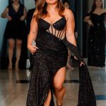 Woman in a black, sparkly gown walks on a red carpet with others in formal attire behind her.