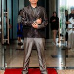 A man in a shiny suit stands on a red carpet in front of glass doors at an indoor event.