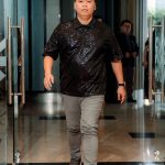Person in a black sequin shirt and gray pants walking on a red carpet indoors.