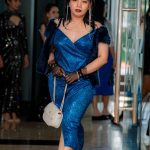 Woman in a blue sparkly dress walking indoors, holding a white purse and wearing black gloves.
