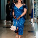 Woman in a blue dress walking on a red carpet indoors, holding a white purse and smiling.