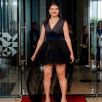 Woman in a black dress stands on a red carpet inside a building, smiling at the camera.