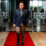 A man in a suit walks on a red carpet through glass doors at an indoor event.