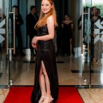 Woman in a black evening gown stands on a red carpet in a formal indoor setting.