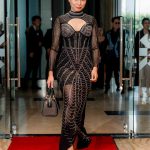Woman in a black, beaded gown walks on a red carpet, holding a handbag.