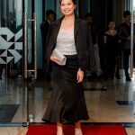 Woman in a black skirt suit and silver top walking on a red carpet, holding a clutch.