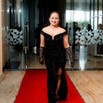 Woman in a black evening gown walks on a red carpet in a modern indoor setting.