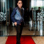 Man in a blue patterned blazer and sunglasses stands on a red carpet by glass doors.