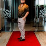 Man standing on a red carpet in front of glass doors, wearing a brown shirt and light pants.
