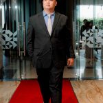 Man in a suit and sneakers walks on a red carpet in a modern building lobby.