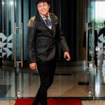 A person in formal attire walks on a red carpet through glass doors, smiling.