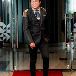 Man in a black suit with gold shoulder accents standing on a red carpet indoors.