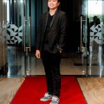 Person in a black suit and sneakers standing on a red carpet in front of glass doors.