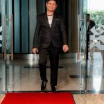 A man in a black suit walks onto a red carpet through glass doors.