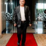 Man in a black blazer and white shirt walking on a red carpet in a modern building.