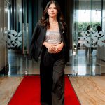 Woman in a black suit and sequin top walks on a red carpet in a modern building lobby.