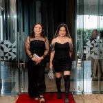 Two women in black outfits walk together on a red carpet through glass doors indoors.