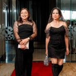 Two women in black dresses walk on a red carpet inside a building with glass doors.