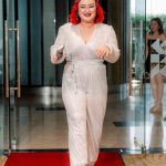 Person in a white sparkly jumpsuit and red heels walks on a red carpet through glass doors.