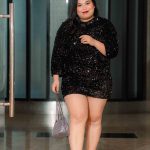 Woman in a black sequin dress and clear heels holding a silver purse, walking indoors.