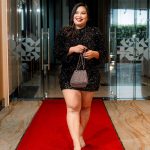 Woman in a black dress walks on a red carpet holding a small purse indoors.